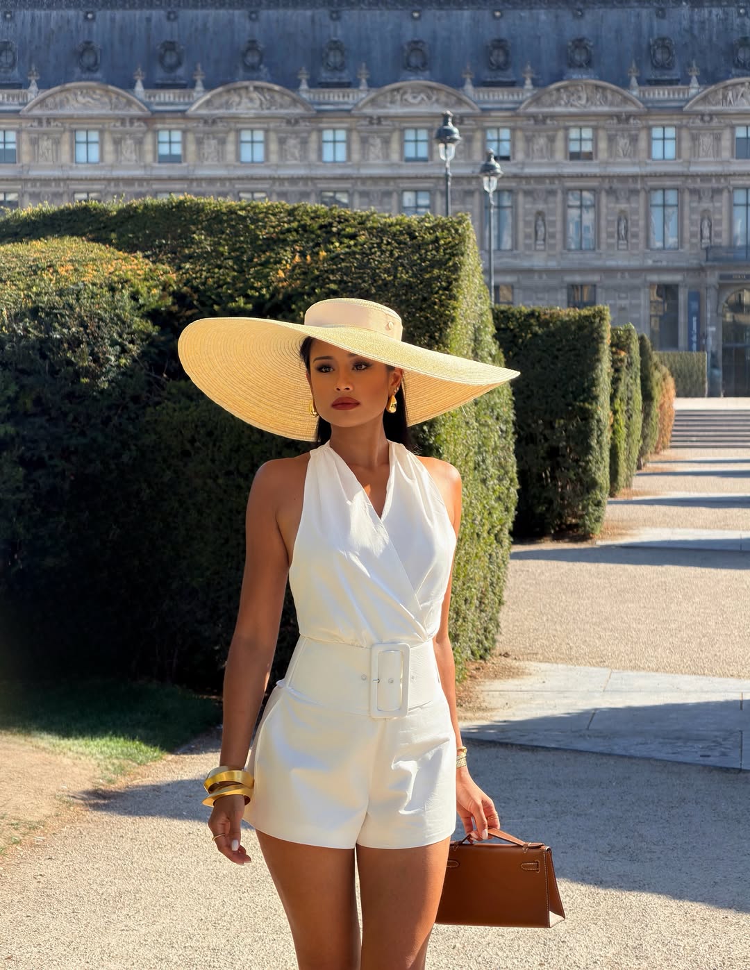 Pat Chaveesak in a white short jumpsuit and wide brimmed straw hat in the Tuileries gardens, the Louvre palace behind her, full length shot in hard summer daylight, self shot